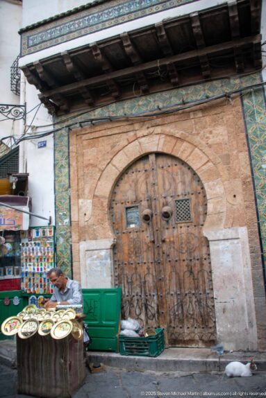 Weathered studded wooden double door in a broad stone horseshoe arch, with green tile borders and a street display of ceramic plates.