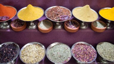 Bowls of yellow and tan powders, red flakes, dried flowers, seeds, and green herbs arranged on a burgundy cloth.