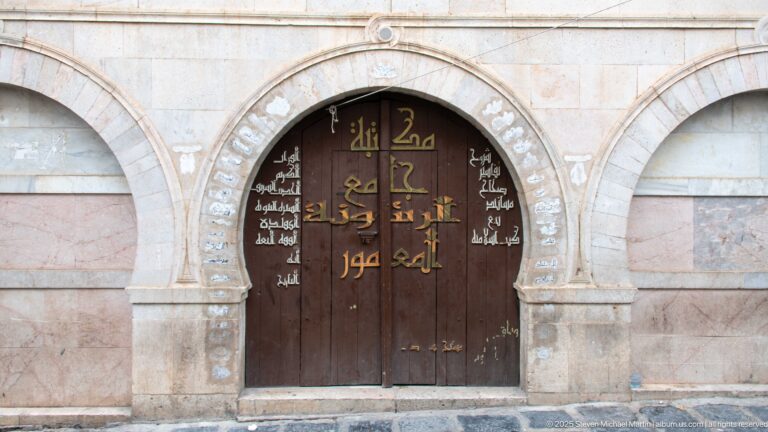 Dark wooden arched double door bearing large gold Arabic lettering and smaller white Arabic script within a pale stone façade.