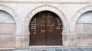 Dark wooden arched double door bearing large gold Arabic lettering and smaller white Arabic script within a pale stone façade.