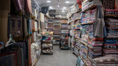 Narrow textile shop with floor-to-ceiling stacks of folded blankets, rugs, towels, and hanging woven goods along a central aisle.
