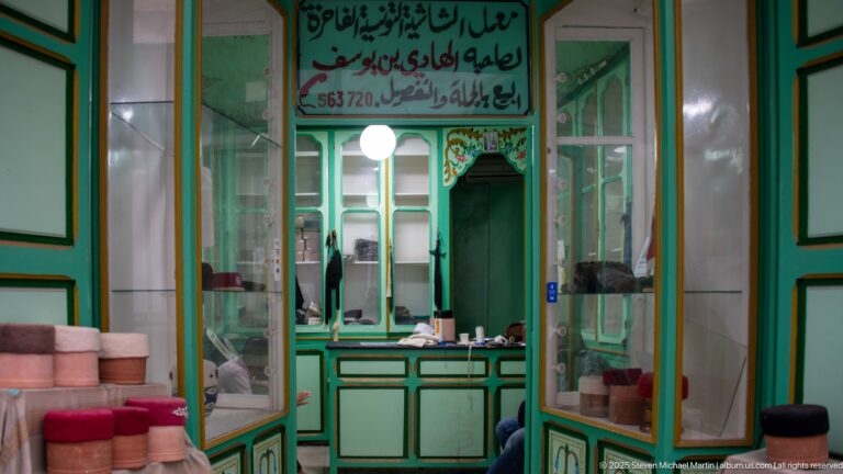 Pale green shop interior with glass cabinets, counter, mirrors, stacked cylindrical goods, and Arabic signage above the service area.