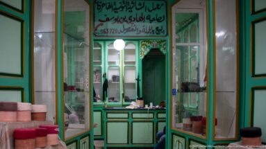 Pale green shop interior with glass cabinets, counter, mirrors, stacked cylindrical goods, and Arabic signage above the service area.