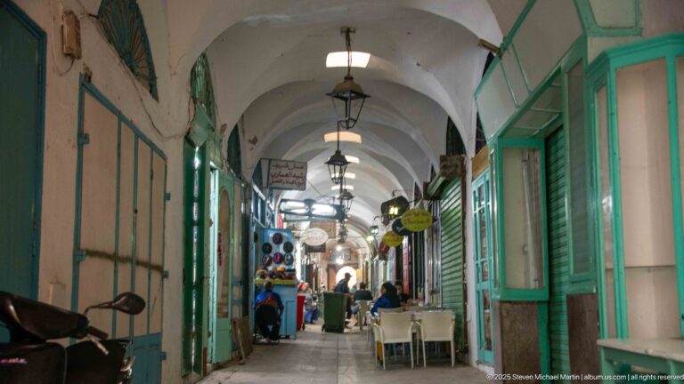 Long vaulted souk passage with hanging lanterns, turquoise shopfronts, café tables, and pedestrians receding toward a brighter opening.