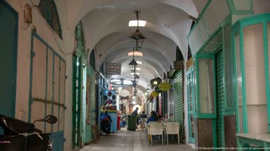 Long vaulted souk passage with hanging lanterns, turquoise shopfronts, café tables, and pedestrians receding toward a brighter opening.