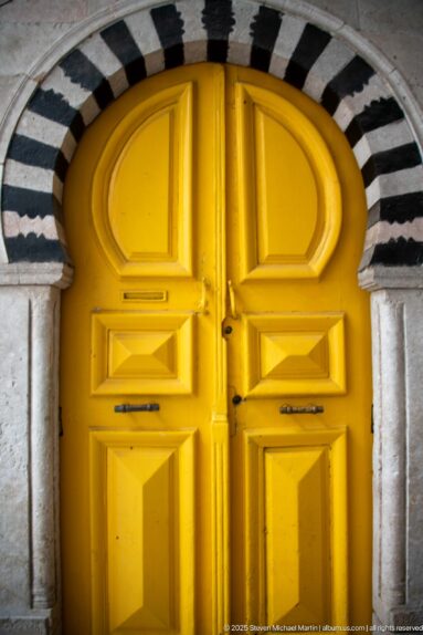 Bright yellow paneled double door beneath an arch of alternating dark and light stone bands.