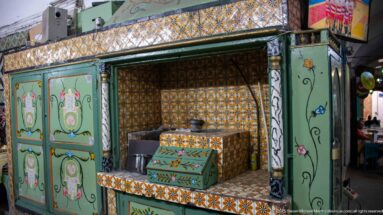 Green-painted kiosk or counter with floral panels and dense multicolored tilework, with a metal pot resting behind the work surface.