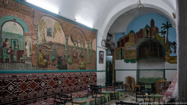 Vaulted room with large painted murals, patterned cushions and textiles, and small painted tables and chairs arranged along the walls.