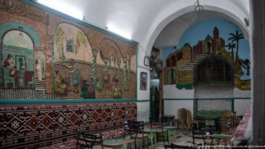 Vaulted room with large painted murals, patterned cushions and textiles, and small painted tables and chairs arranged along the walls.