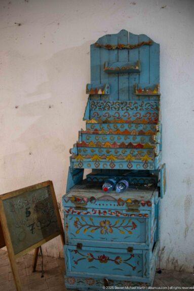 Blue stepped wooden chest with hinged compartments, painted floral and geometric motifs, and worn surfaces against a plain wall.
