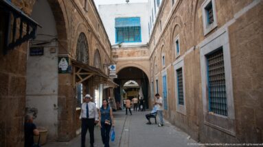 Narrow stone-lined medina lane with blue windows and grilles, pedestrians, shop awnings, and an arched passage crossing the street ahead.