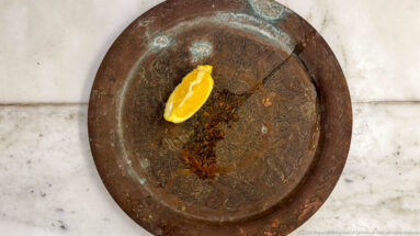 Round engraved copper-toned tray with scrolling patterns, areas of green oxidation, and a bright citrus wedge resting near the center.