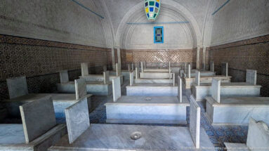 Long mausoleum chamber with rows of raised marble tombs, inscribed headstones, patterned tile walls, carved plaster, and a colored hanging lantern.