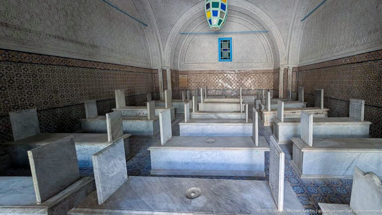 Long mausoleum chamber with rows of raised marble tombs, inscribed headstones, patterned tile walls, carved plaster, and a colored hanging lantern.