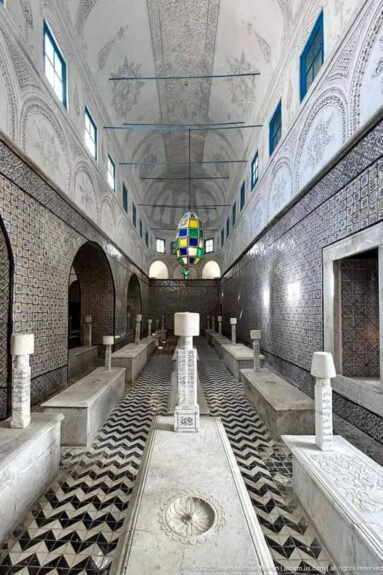 Symmetrical mausoleum hall with rows of marble tombs, black-and-white geometric flooring, tiled walls, carved plaster vaults, and a colored lantern.