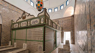 Green latticed funerary enclosure with Arabic calligraphic panels, surrounded by marble tomb markers in a richly tiled chamber.
