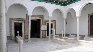 White arcaded courtyard with slender columns, raised marble tombs, patterned tile borders, dark doorways, and green roof trim.