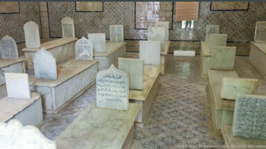 Crowded mausoleum chamber with raised marble tombs, upright Arabic-inscribed headstones, patterned floor tiles, and richly tiled walls.