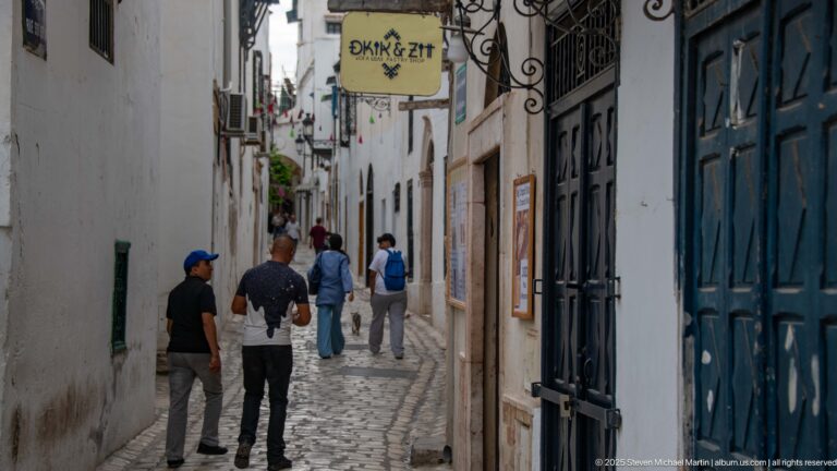 Pedestrians walking along a narrow whitewashed medina lane with dark doors, wrought-iron brackets, and hanging shop signs.