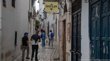 Pedestrians walking along a narrow whitewashed medina lane with dark doors, wrought-iron brackets, and hanging shop signs.