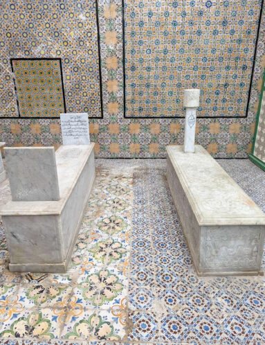 Two raised marble tombs with upright memorial markers on a richly patterned tiled floor against floral and geometric tile-covered walls.