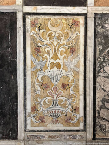 Rectangular polychrome stone inlay panel with a symmetrical vase, scrolling foliage, and stylized flowers framed by pale stone borders.