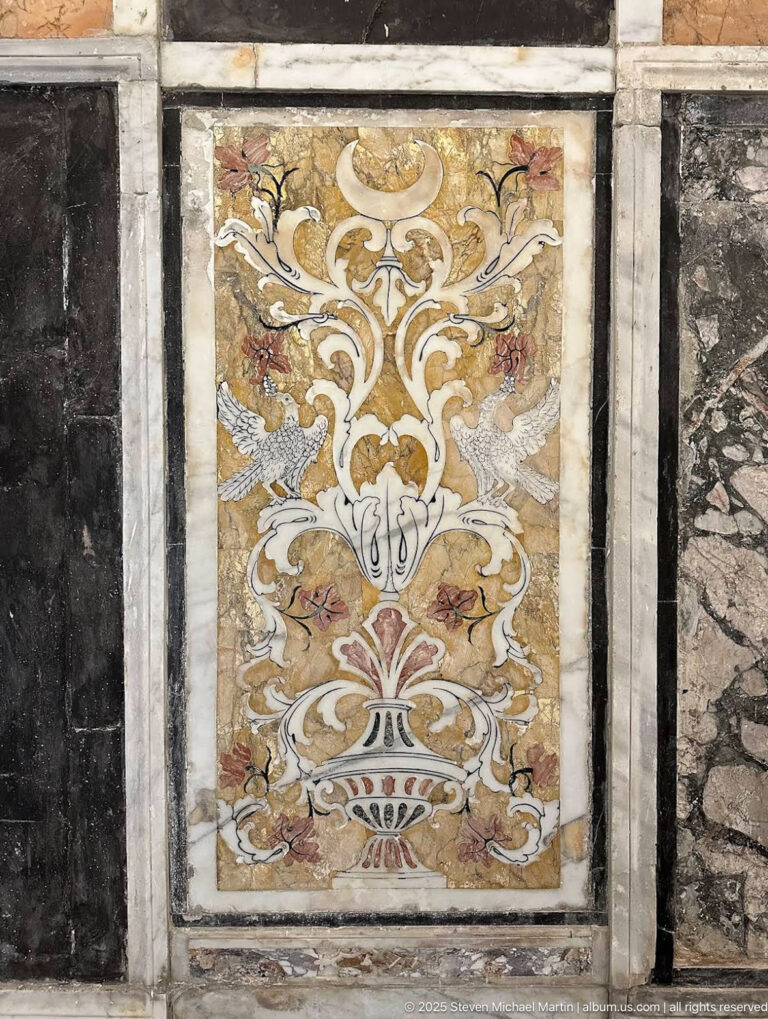 Rectangular polychrome stone inlay panel with a symmetrical vase, scrolling foliage, and stylized flowers framed by pale stone borders.