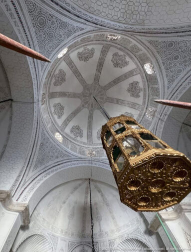 Upward view of an intricately carved white plaster vault with radial ornament and a large gilded hanging lantern in the foreground.