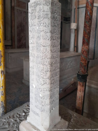 Pale rectangular interior pillar covered with multiple lines of Arabic script, beside carved surfaces and a reddish support post.