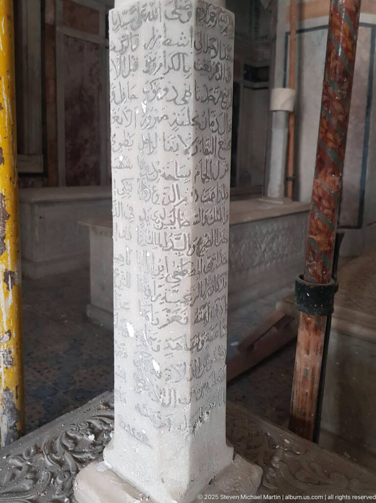 Pale rectangular interior pillar covered with multiple lines of Arabic script, beside carved surfaces and a reddish support post.