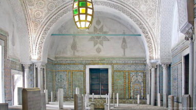 Mausoleum chamber with raised marble tombs, richly tiled walls, carved white plaster vaults, stone columns, and a multicolored hanging lantern.