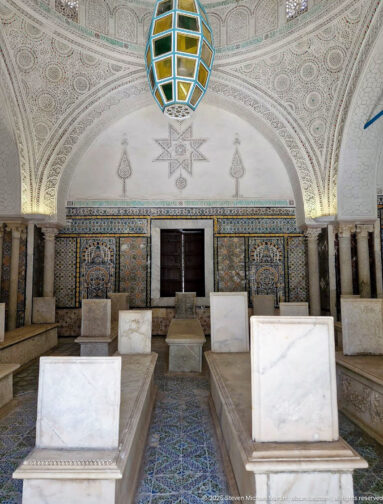 Rows of marble tombs aligned toward a dark doorway in a richly tiled mausoleum chamber beneath carved plaster vaults and a colored lantern.