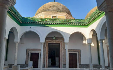 White arcaded mausoleum courtyard with slender columns, marble tombs, green-tiled cornice, and a large patterned dome rising above.
