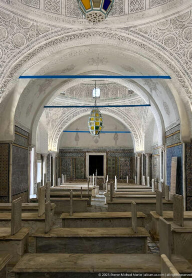 Wide mausoleum interior with repeated white arches, rows of marble tombs, tiled walls, carved plaster vaults, columns, and a colored lantern.