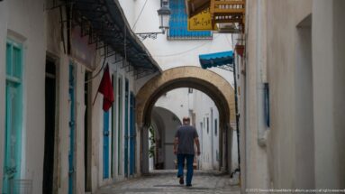 Man walking toward successive masonry arches on a narrow medina lane with turquoise doors, signs, and a Tunisian flag.