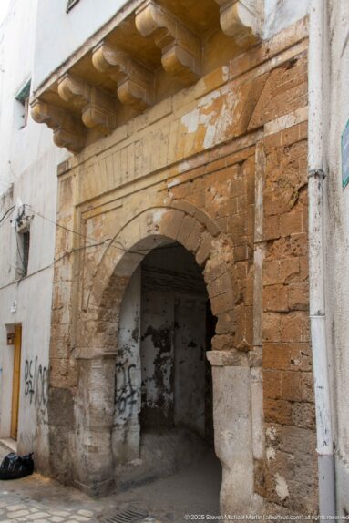 Weathered stone-and-brick arch spanning a shallow medina recess, with worn plaster, masonry piers, and graffiti.