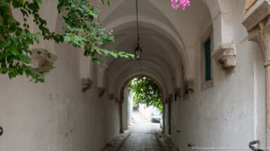 White ribbed medina passage with a hanging lantern, stone corbels, and bougainvillea framing the bright exit.