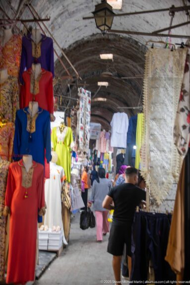 Covered souk aisle beneath a worn vault, lined with colorful clothing displays and shoppers moving through the narrow passage.