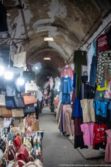 Long vaulted souk aisle with piles of shoes, hanging handbags, clothing racks, and bright electric lights.