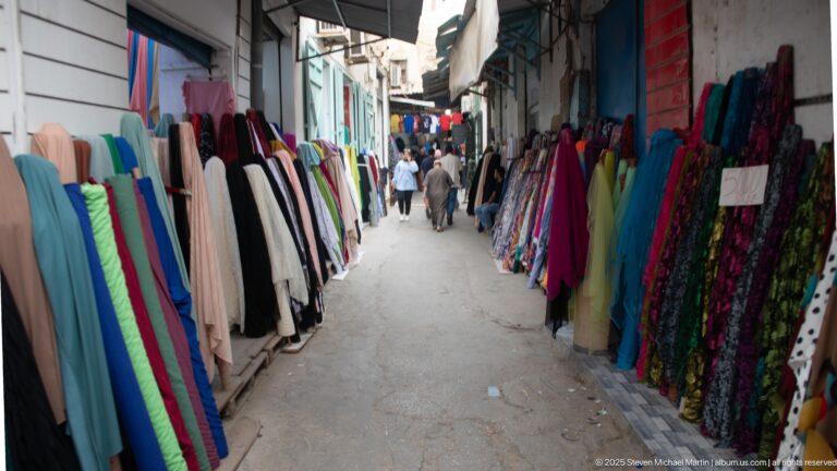 Narrow souk lane lined on both sides with upright bolts of colorful fabric as pedestrians walk through the center.