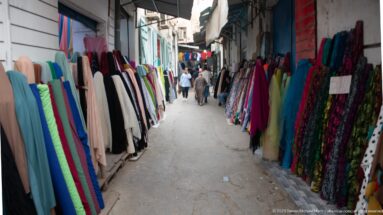 Narrow souk lane lined on both sides with upright bolts of colorful fabric as pedestrians walk through the center.