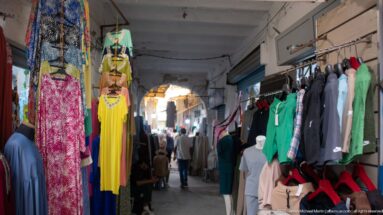 Covered medina market passage with dresses and clothing racks on both sides and shoppers beneath an arch in the distance.