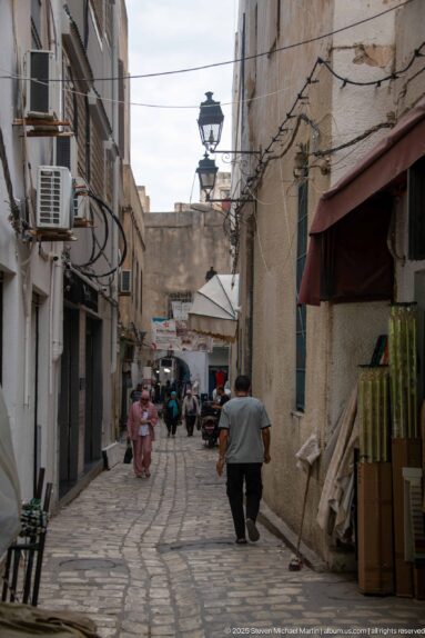 Cobbled medina commercial lane with pedestrians, shop awnings, exposed wiring, air-conditioning units, and stacked goods.
