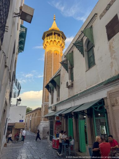 Octagonal masonry minaret with a projecting decorated gallery rising above white medina buildings with green shutters and shopfronts.