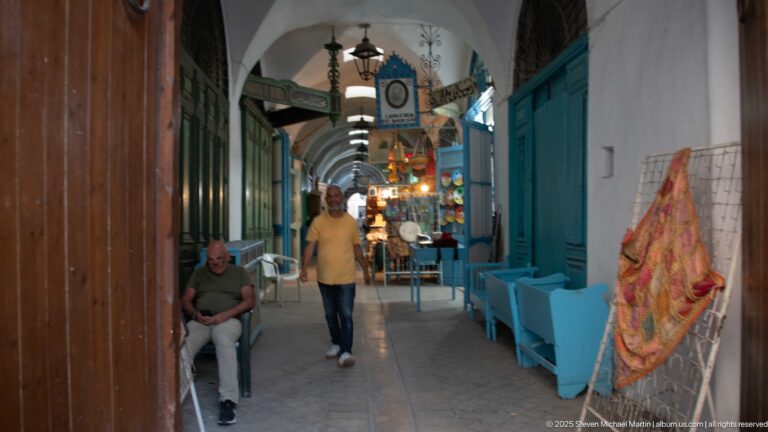 Narrow vaulted souk passage with turquoise shopfronts, benches, hanging lamps, signs, and pedestrians.