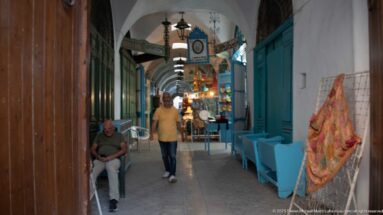 Narrow vaulted souk passage with turquoise shopfronts, benches, hanging lamps, signs, and pedestrians.