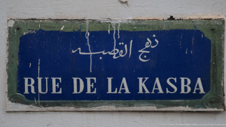 Weathered blue street plaque reading “RUE DE LA KASBA” beneath Arabic script on a white wall.