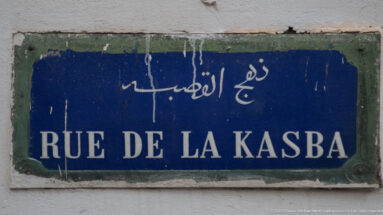 Weathered blue street plaque reading “RUE DE LA KASBA” beneath Arabic script on a white wall.