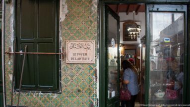 Entrance to Le Foyer de l’Artiste with bilingual sign, green shutters, patterned tilework, and a visitor entering the shop.