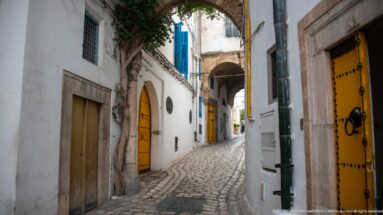 Narrow cobbled medina alley lined with yellow doors and white walls, leading beneath an overhead arch toward a covered passage.